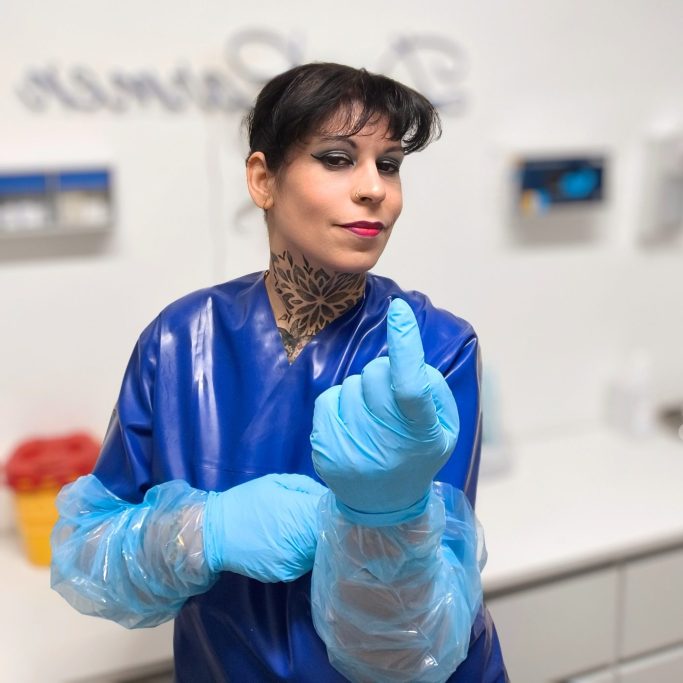 Frau Doktor Carmen Frau mit blauen Handschuhen und Kleidung zeigt Daumen nach oben in einem klinischen Umfeld.